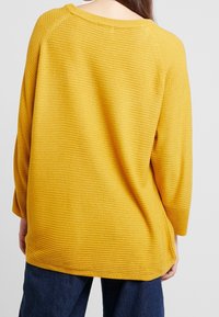 Musztardowożółty sweter o fakturowanym, prążkowanym wzorze i rękawach do łokcia. Posiada szeroki dekolt i luźny krój.
