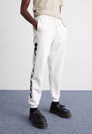 Hvide joggers med et sort grafisk design langs venstre ben, udformet med elastisk talje og sidelommer. Sorte sko fuldender looket.