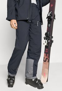Μπλε ναυτικό παντελόνι χιονιού με γκρι λεπτομέρειες, αδιάβροχο υλικό, ρυθμιζόμενη μέση και τσέπες με φερμουάρ, σε συνδυασμό με μαύρα παπούτσια σκι και σκι.