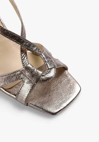 Metallisk bronze sandal med krydset remme, firkantet tå og en glat overflade. Har en subtil glans og et minimalistisk design.