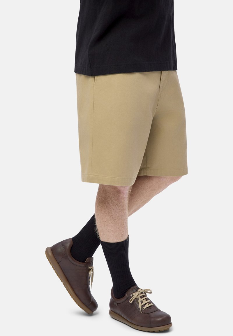 Person wearing beige shorts, black socks, brown sneakers with beige laces, and a black shirt visible from waist down, standing on one leg.