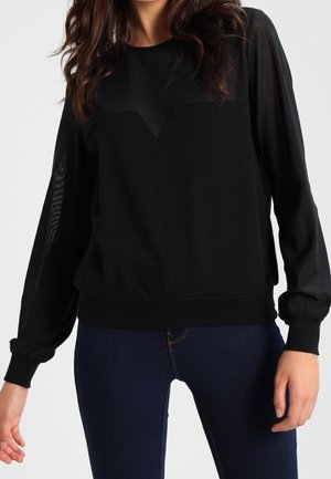 Kvinde iført en sort langærmet top med gennemsigtig panel og mørkeblå jeans, stående med den ene arm let bøjet.
