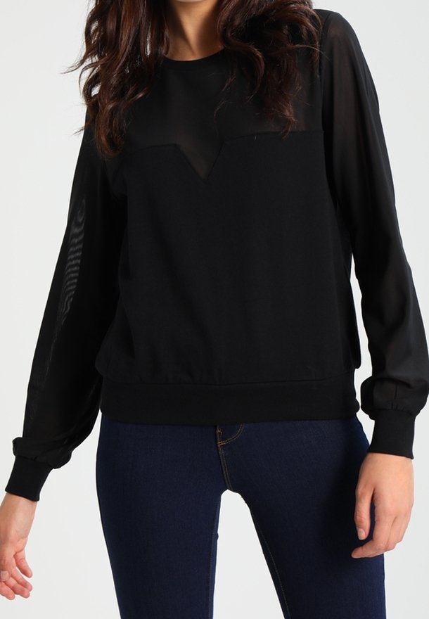 Femme portant un haut noir à manches longues avec panneau transparent et un jean bleu foncé, debout avec un bras légèrement plié.