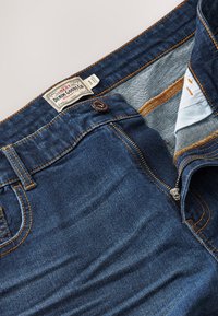 Dark blue denim jeans with partially open zipper, visible copper button, orange stitching, and a white brand label inside the waistband.