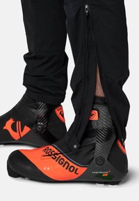Schwarze Skischuhe mit orangen Akzenten, Karbonfasertextur und einem Reißverschlussmerkmal an der Hose. Der Schuh trägt den Markennamen Rossignol.