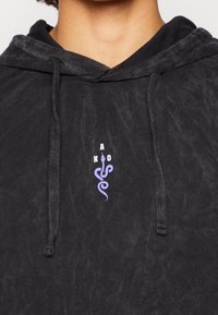 Sort hættetrøje med tekstureret finish, der har en hvid og lilla slangelogo samt bogstaverne 'A K O' på forsiden. Justerbar snøre på hætten.