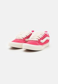 Paire de baskets basses rouges et blanches avec des lacets blancs sur un fond blanc uni.