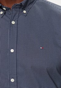 Camisa azul marino de botones con un patrón cuadrado blanco, con cuello, botones blancos y un pequeño logotipo en rojo, blanco y azul.