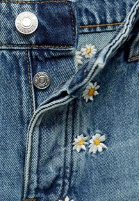 Jeans tyg med en ljusblå tvätt. Har silverfärgade metallknappar och broderade vita tusenskönor med gula mittar längs sömmen.