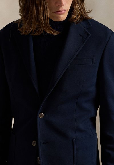 Person with shoulder-length hair wearing a dark navy blazer over a black top, shown from the chest up against a neutral background.