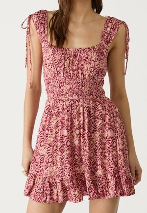 Femme portant une robe sans manches rose à motifs avec bretelles à nouer, corsage froncé et ourlet à volants sur un fond uni.