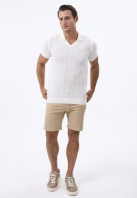 Polo de punto blanco de manga corta con cuello en V, combinado con pantalones cortos de lino beige y zapatillas beige, con un patrón texturizado.