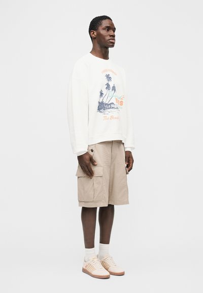 Άνδρας που στέκεται φορώντας λευκό φούτερ με σχέδιο φοίνικα, μπεζ cargo σορτς, λευκές κάλτσες και μπεζ αθλητικά παπούτσια, μπροστά σε απλό φόντο.