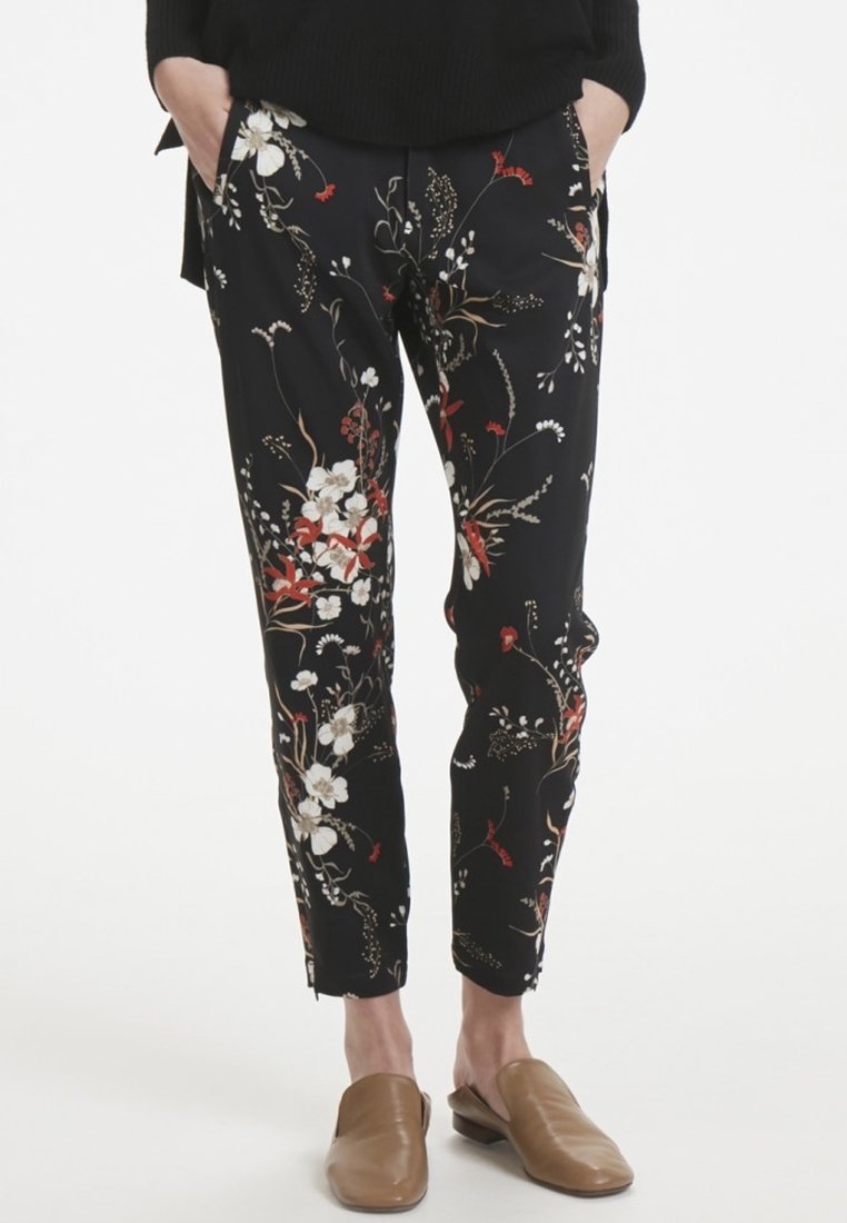 Pantalon noir à imprimé floral avec des fleurs blanches et rouges. Design ajusté avec poches latérales et coupe à la cheville. Texture de tissu lisse.