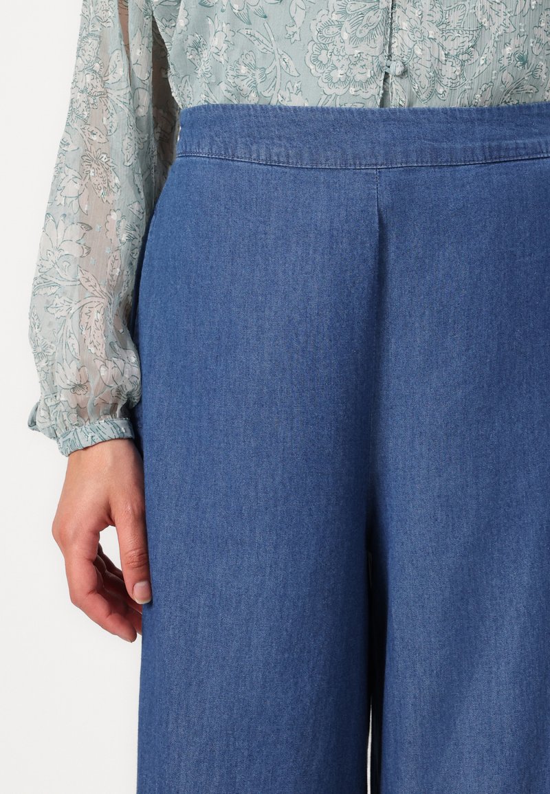 Pantaloni ampi in denim azzurro chiaro, con vita alta, trama sottile e cuciture lisce. Abbinati a una blusa trasparente con motivo floreale.