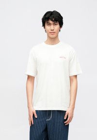 Hvid t-shirt i bomuld med korte ærmer, med en lille rød broderet tekst på brystet. Kombineret med blå bukser med fine liner.