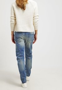 Vit stickad tröja med långa ärmar; blå denimjeans med en blekt finish och synliga sömmar; slitna vita sneakers som fulländar outfiten.