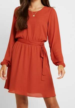 Femme portant une robe à manches longues couleur rouille avec une ceinture à la taille et un collier pendentif en or, debout devant un fond uni.