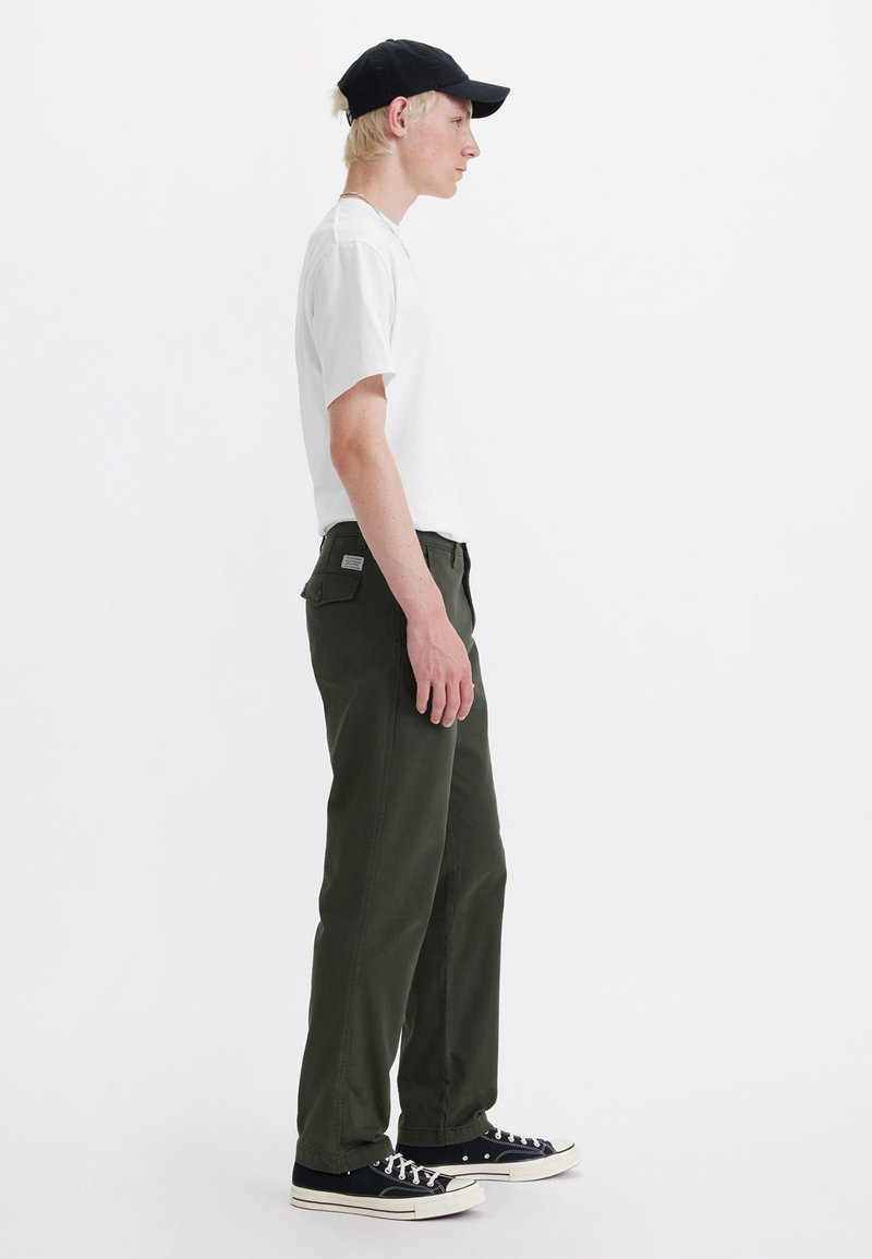 Conjunto casual que incluye una camisa blanca de manga corta, pantalones verde oscuro con un bolsillo trasero y zapatillas negras con detalles blancos.