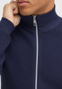 Námornícka modrá textúrovaná šušťáková bunda so zipsom a vysokým golierom, s bielym zipsom. Materiál má vystúpený vzor, ktorý pridáva hĺbku dizajnu.