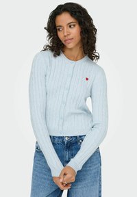 Gaiši zils kabeļu adījuma kardigans ar apaļu izgriezumu, pogu aizdari un nelielu sarkanu sirds emblēmu kreisajā krūšu daļā.