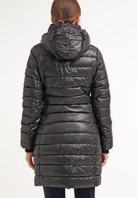 Žena ve dlouhém černém prošívaném péřovém kabátu s kapucí, stojící zády k jednoduchému bílému pozadí.