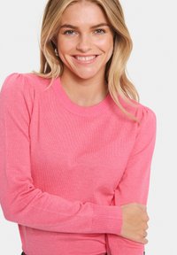 Roza pletena jopica z okroglo izrezom, rebrastimi manšetami in rahlimi napihnjenimi rokavi. Mehka tekstura in enakomeren vzorec šivanja po celotni dolžini.