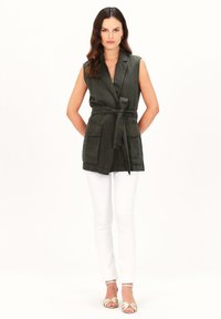 Mouwloze donkergroene vest met een strik taille, twee voorkanten zakken en een kraag, gecombineerd met witte skinny broek en lichtgekleurde sandalen.