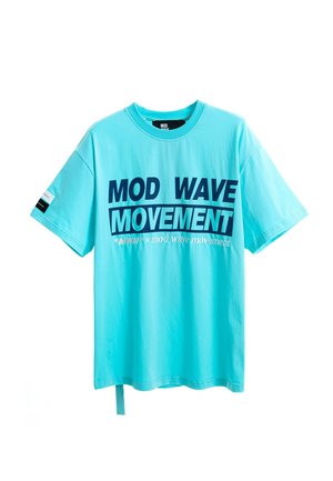 Joven de pie con una camiseta turquesa con la inscripción "MOD WAVE MOVEMENT", pantalones cortos vaqueros azules, calcetines blancos con diseño negro y zapatos beige texturizados sin cordones.