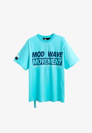 Joven de pie con una camiseta turquesa con la inscripción "MOD WAVE MOVEMENT", pantalones cortos vaqueros azules, calcetines blancos con diseño negro y zapatos beige texturizados sin cordones.