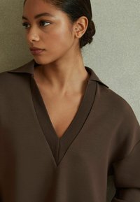 Maglione marrone a V con vestibilità rilassata, colletto a coste e texture liscia, caratterizzato da spalle scese e un design semplice e pulito.