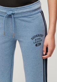 Jasnoniebieskie dresy z fakturowanym wykończeniem, z czarnym nadrukiem "Superday Athl League 82" oraz kontrastującymi czarnymi bocznymi paskami.