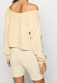 Ljusbeige kort sweatshirt med off-the-shoulder-design och långa ärmar, matchad med texturerade beigefärgade shorts.