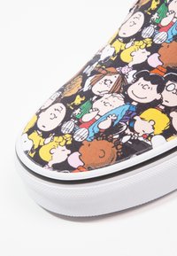 Baskets avec une tige en tissu noir présentant un motif cartoon coloré de personnages. Semelle en caoutchouc blanc avec une surface texturée et une bordure noire.