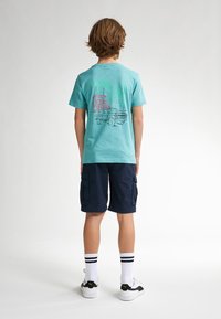 Tyrkysové bavlněné tričko se zadním potiskem vintage náklaďáku a budov, kombinované s námořnickými cargo šortkami a bílými pruhovanými ponožkami.