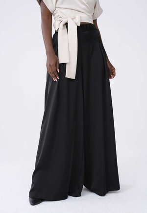 Femme portant un pantalon noir taille haute à jambes larges et un top cache-cœur crème noué en nœud à la taille, debout sur fond blanc.