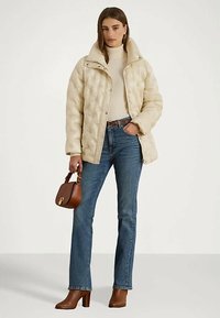 Moteris vilki rusvą siūlėtų paltą, kremine megztinė, mėlynus džinsus, rudas aulinukus, laikydama rudą rankinę su trumpomis rankenomis.