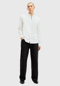 Hvid langærmet skjorte med knapper og afslappet pasform, parret med sorte straight-leg jeans og sorte sko, mod en neutral baggrund.