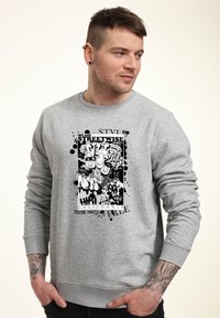Muž s tetováním, který má na sobě šedou mikinu s černobílým graffiti-style grafickým vzorem a textem "Jsme chytří z ulice Kalifornie."