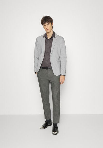 Blazer cinza claro, camisa de botão cinza escuro, calças cinzas com padrão e sapatos pretos com detalhes em prata. Corte ajustado.