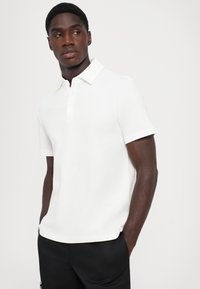 Hvit kortermet poloskjorte med teksturert overflate, tre-knapps placket og lett buet kant, parret med svarte shorts.