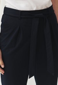 Pantaloni blu navy con stringa in vita, presentano pences sulla parte anteriore e una texture del tessuto liscia. Senza tasche visibili.