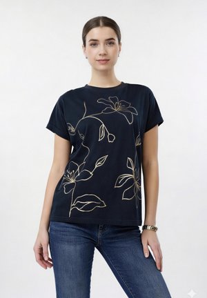 Junge Frau trägt ein schwarzes T-Shirt mit goldenem Blumendesign und blaue Jeans, steht vor einem schlichten, hellen Hintergrund.
