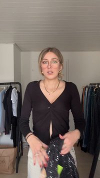 Must meisterdatud must ülaosa, millel on keerutatud esi detail, pikad varrukad ja sügav V-kaelus. Taustal on riideid riidepuudel ja kott.