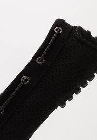 Gros plan sur le talon d'une chaussure en daim noir avec un motif en nid d'abeille, des lacets tressés passant à travers des œillets métalliques, et des picots en caoutchouc sur la semelle.