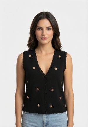 Femme aux longs cheveux foncés porte un top noir sans manches à boutons avec des embellissements floraux dorés et un jean bleu clair, debout sur un fond blanc.