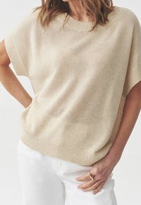 Donna che indossa un top a maglia beige a maniche corte e pantaloni bianchi, con una mano sui fianchi e un anello d'oro al dito.