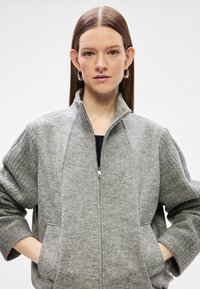 Νεαρή γυναίκα με ίσια καστανά μαλλιά φορά ασημένια σκουλαρίκια κρίκους και ένα γκρι μάλλινο φούτερ με φερμουάρ και ραβδωτά μανίκια, με τα χέρια στις τσέπες.