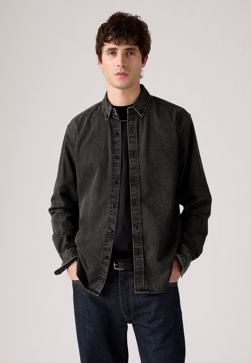 Dark gray denim shirt with button closure, pointed collar, and long sleeves. Worn over a black T-shirt, paired with blue denim pants.