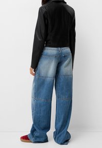 Czarna skórzana kurtka o kroju cropped w połączeniu z niebieskimi spodniami dżinsowymi typu wide-leg z patchworkiem, charakteryzującymi się różnymi odcieniami denimu i oversize'owym krojem. Widoczne czerwone obuwie.